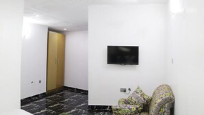 A hotel room with a flat-screen TV mounted on the wall, a floral-patterned chair, and a wooden door. - Volantis Gold (Lagos)