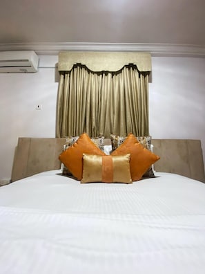 A hotel room with a bed, a headboard, a curtain, and a wall-mounted air conditioning unit. - Volantis Gold (Lagos)