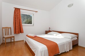 Une chambre avec un lit en bois, une literie blanche, un plaid orange, une chaise en bois, une table de chevet avec une lampe et une fenêtre avec des rideaux.