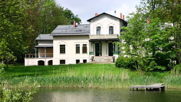 A two-story house with a balcony and a dock extending into a pond. - Seminar & vacation apartment - Gutshaus Bartelshagen (Dalkendorf)