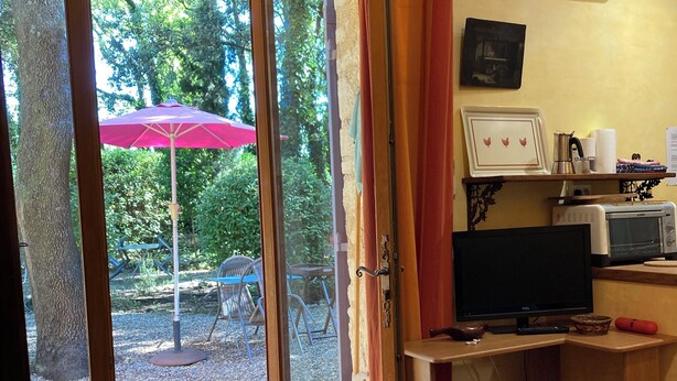 A room with a wooden floor, a sliding glass door leading to an outdoor area with a red umbrella, a TV, a microwave, and a shelf with various items.