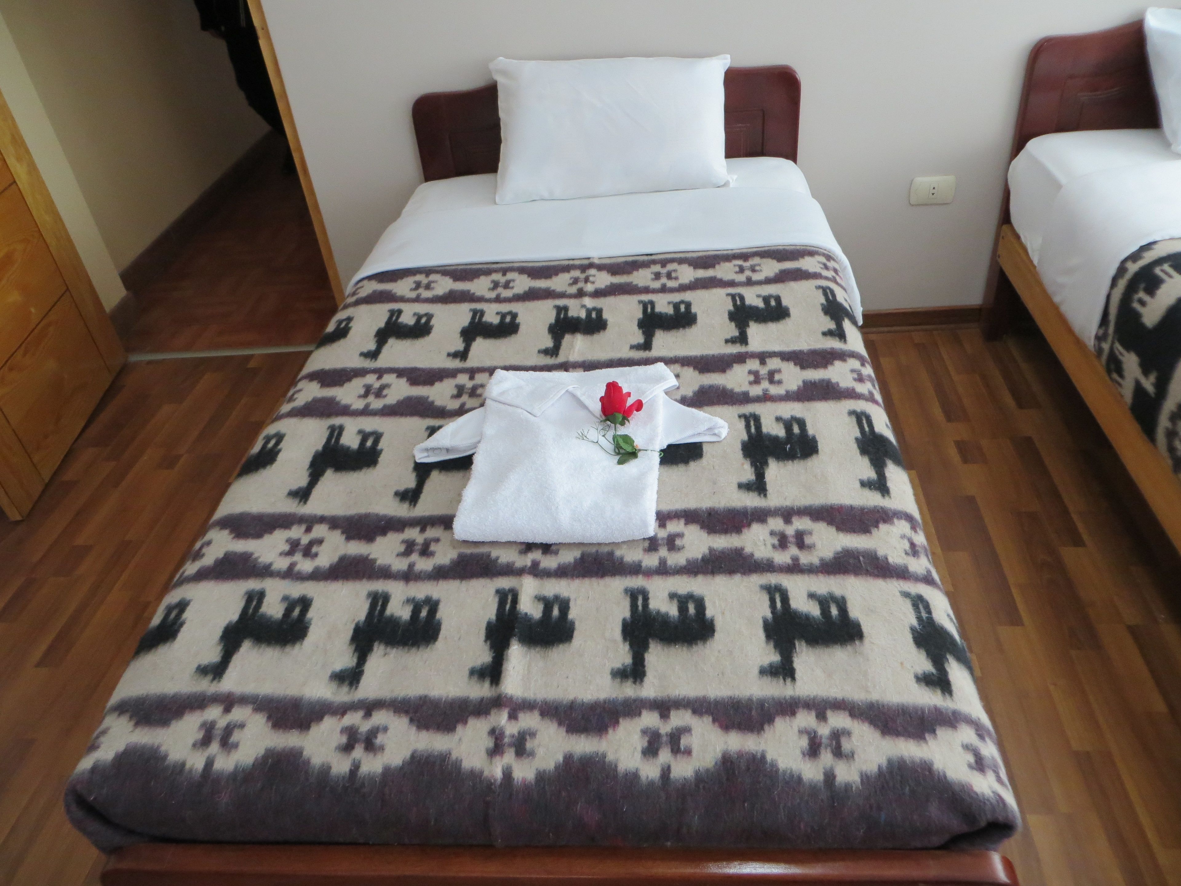 Una cama bien hecha con un edredón estampado, una almohada blanca y una toalla blanca doblada con una rosa roja.