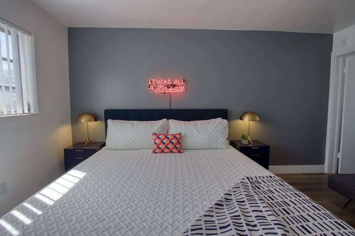 A neatly made bed with a patterned blanket, two bedside tables with lamps, a neon sign on the wall, and a window with blinds.