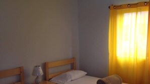 Une chambre avec deux lits simples, chacun doté d’une tête de lit en bois et d’un matelas blanc. Une table de chevet avec une lampe se trouve de chaque côté de chaque lit. Une serviette roulée est posée sur le lit, devant la fenêtre, qui est ornée de rideaux jaunes.