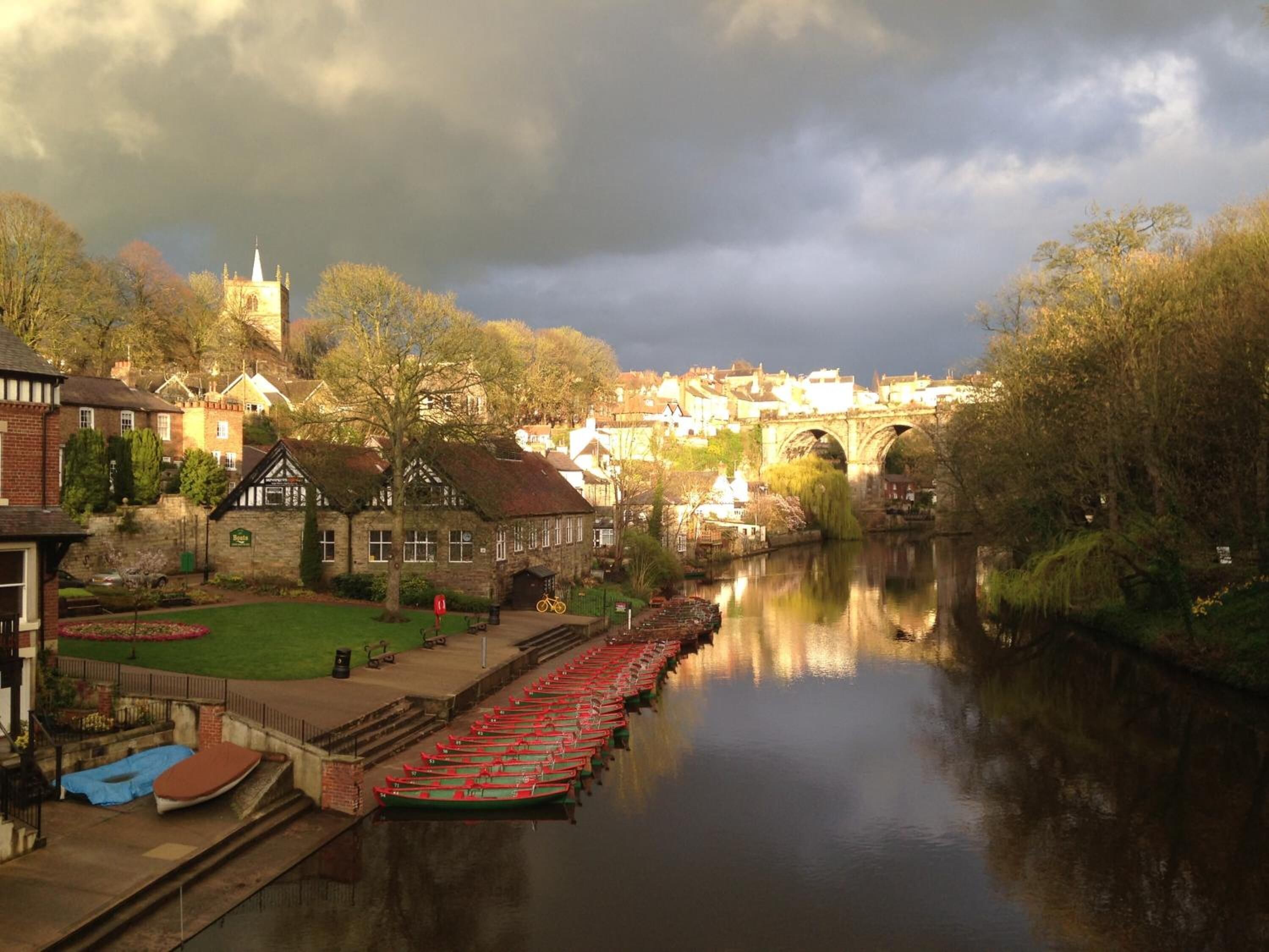 A riverside town with a row of boats docked along the water, a historic bridge, and traditional buildings.