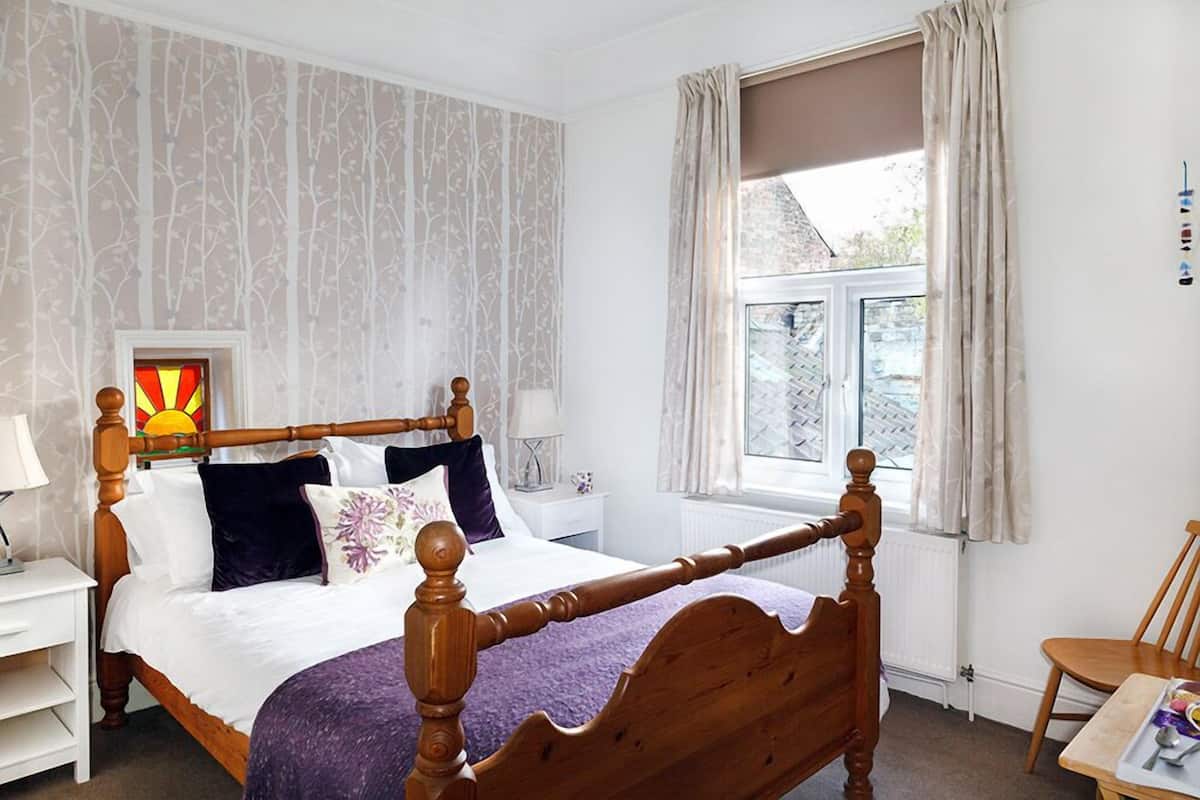 A bedroom with a wooden four-poster bed, a white nightstand, a bedside table, a window with curtains, and a view of the outside.
