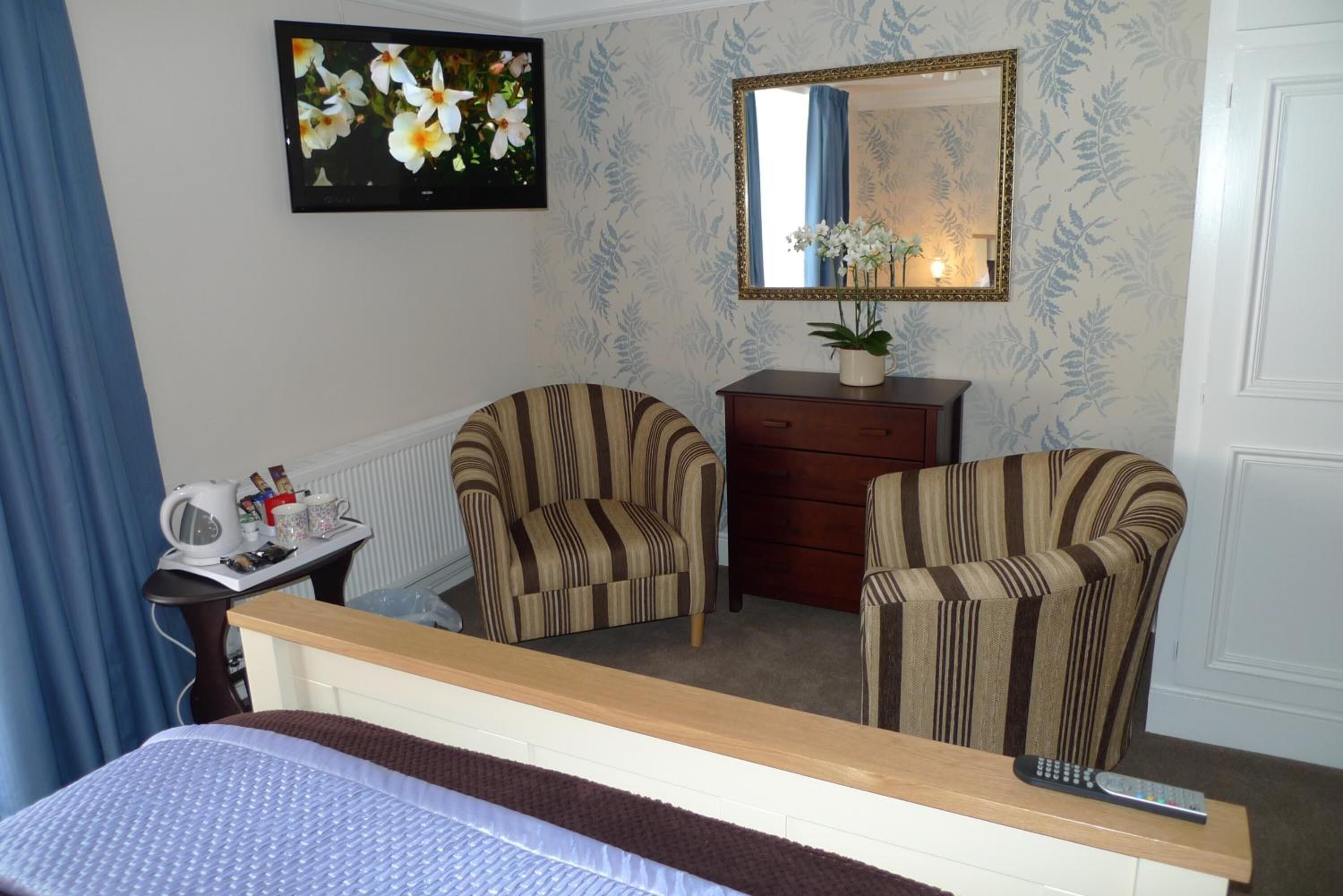 A hotel room with a bed, two striped armchairs, a small table with a kettle and mugs, a dresser, a mirror, and a painting of flowers.