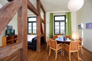 Una habitación con techo de madera, una mesa de comedor con sillas, un televisor en un estante y ventanas grandes con cortinas verdes.