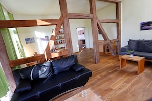 Ein Zimmer mit freiliegenden Holzbalken, einem schwarzen Ledersofa, einem Holztisch und einem Bücherregal.