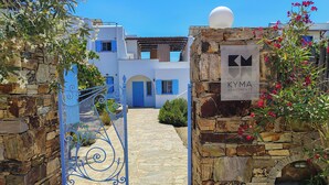 Une porte en pierre surmontée d’une enseigne indiquant « KYMIA APARTMENTS », qui mène à un bâtiment blanc aux volets bleus et à un chemin de pierre.