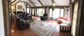 Una sala de estar amplia con un sofá de cuero negro grande, un sillón blanco más pequeño y un sofá rosa. Una alfombra estampada cubre el suelo de madera, y hay un ventilador de techo.