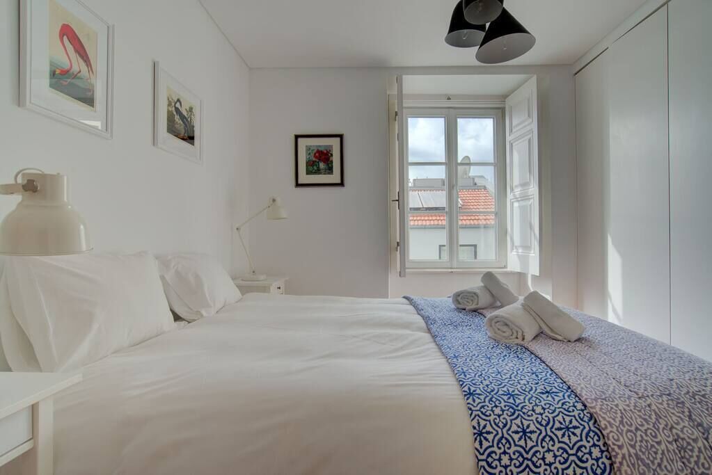 A bedroom with a bed, a bedside table, a lamp, a window with a view of buildings, and a door.