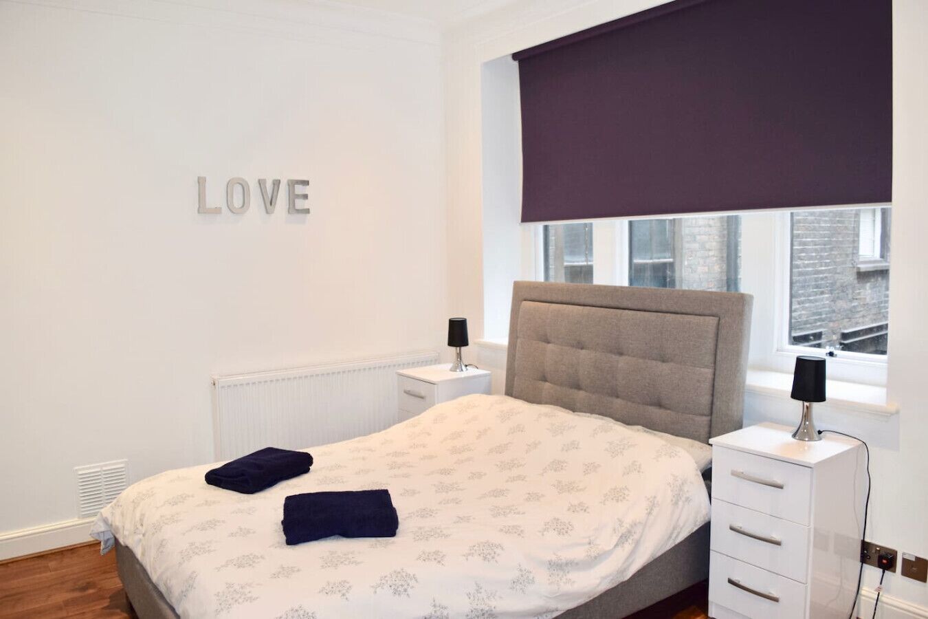 Une chambre à coucher avec un lit, une table de chevet, une lampe, une fenêtre donnant sur un beau paysage et un mur sur lequel est écrit le mot « AMOUR ».
