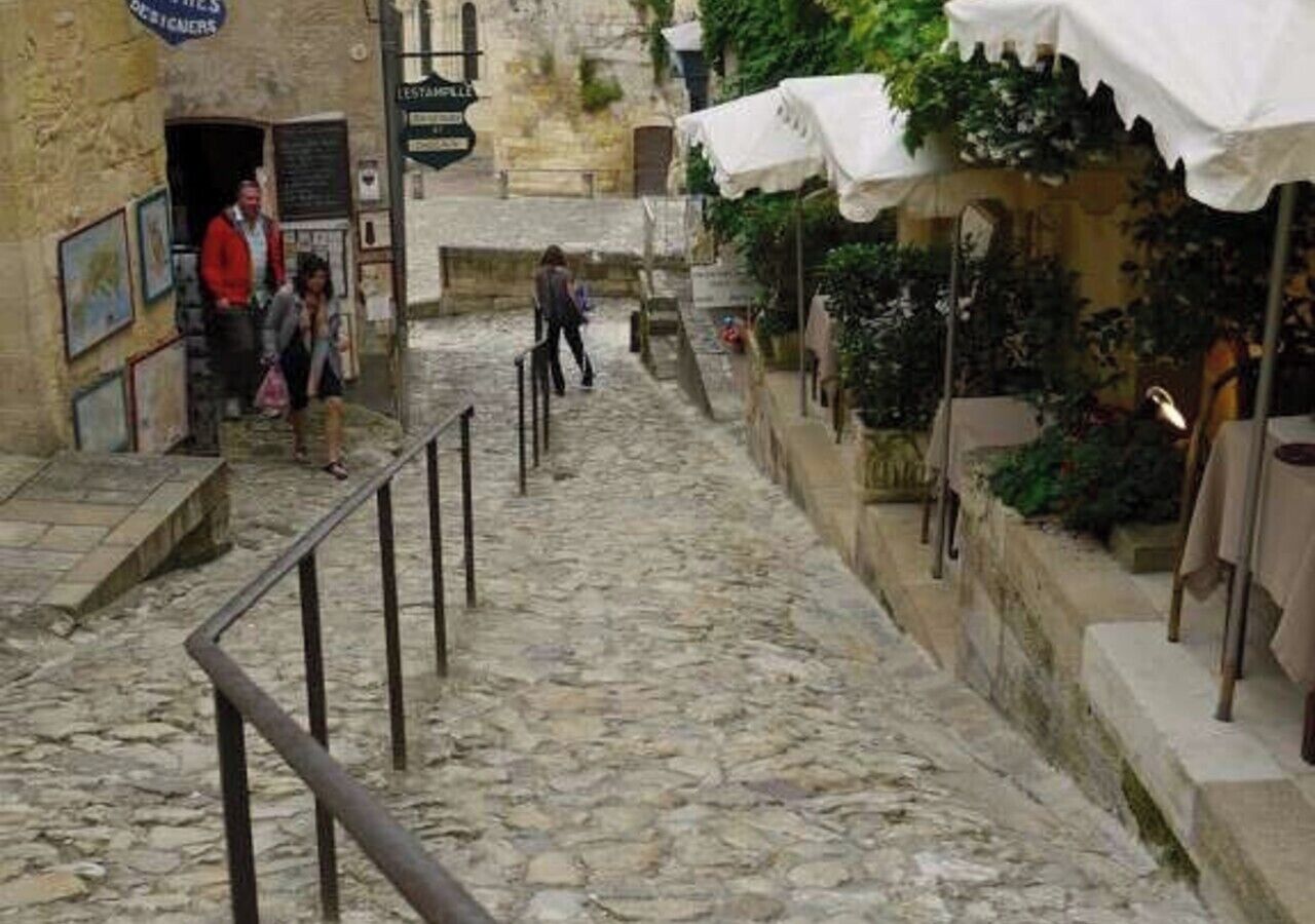Une ruelle étroite pavée, bordée d’une rambarde en métal, avec une entrée d’immeuble et un espace abrité agrémenté de plantes.