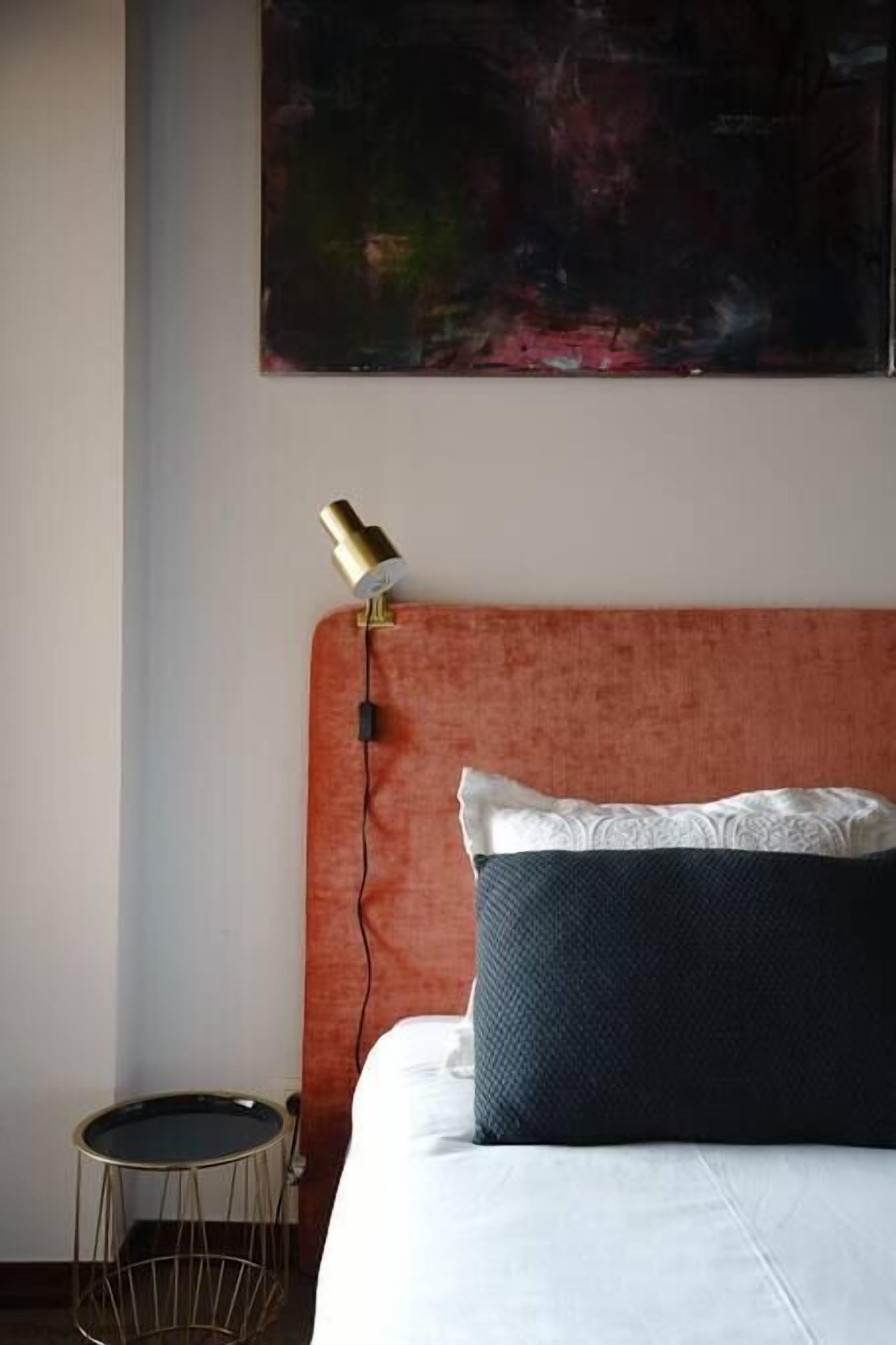 Een slaapkamer met een bed, een rond bijzettafeltje, een lamp en een schilderij aan de muur.