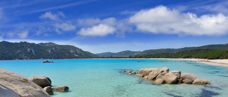 Une mer turquoise aux eaux limpides, avec des rochers visibles au premier plan, une plage de sable et un paysage montagneux en arrière-plan, le tout sous un ciel bleu parsemé de nuages.