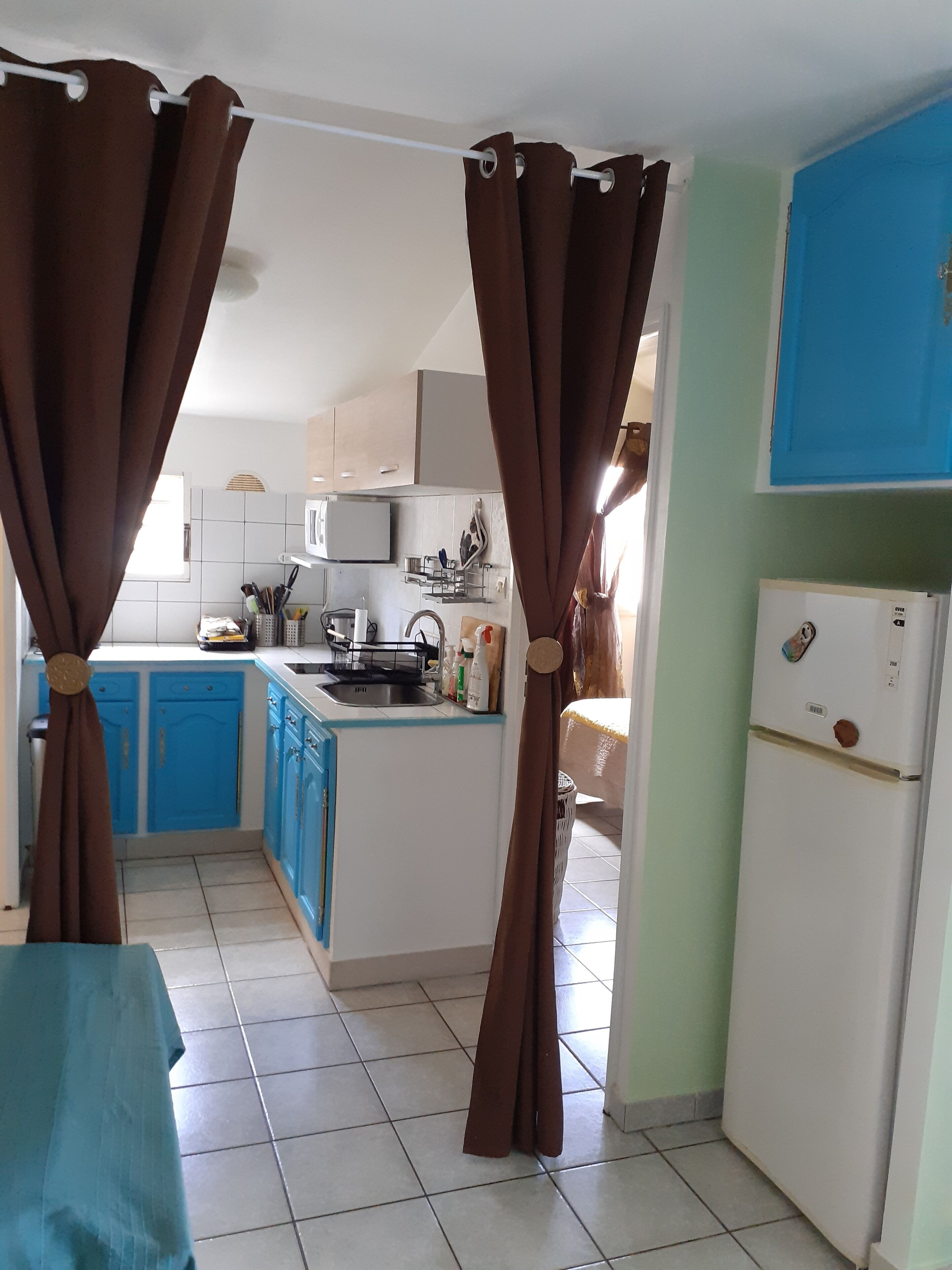 Una cocina compacta con armarios azules, un refrigerador blanco y un microondas. Cortinas marrones atadas en la parte superior.