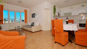 Ett modernt vardagsrum med en soffa, ett matbord med orange stolar, en köksdel och stora fönster med gardiner.
