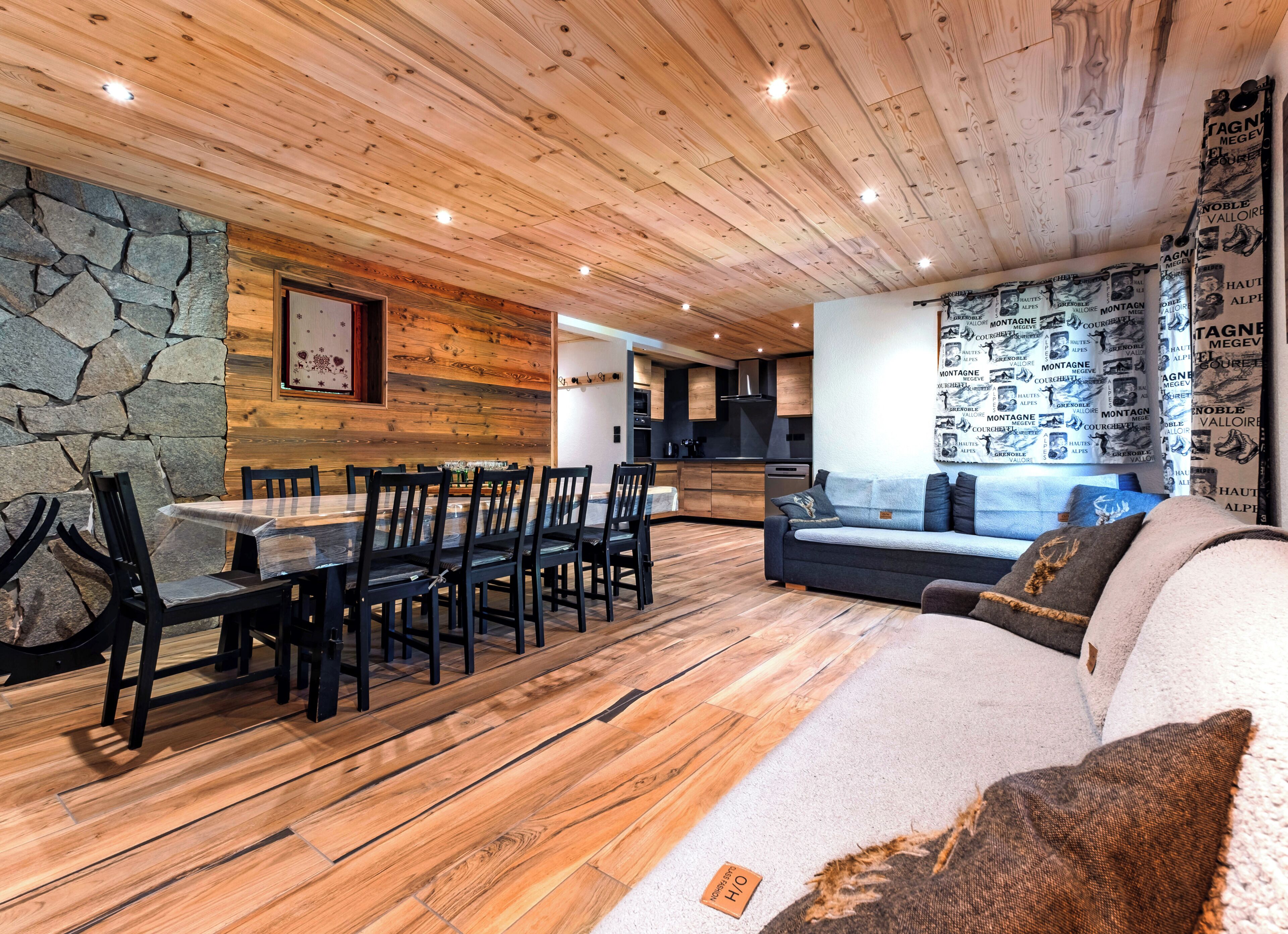 A modern interior with a wooden ceiling, stone wall, dining area with chairs, sofa, and a kitchen in the background.