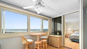Una habitación con un ventanal grande con vistas al océano, un ventilador de techo, una mesa de comedor con sillas y una cama visible a través de una puerta de cristal.