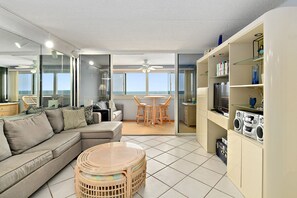 A modern living room with a sofa, a round table, and a TV. There is a balcony with a view of the ocean.