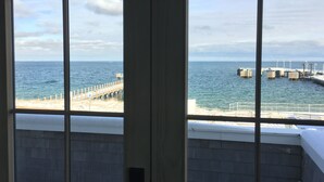 Ein Zimmer mit Meerblick durch große Fenster, eine Holzterrasse und ein Steg mit Bebauung.