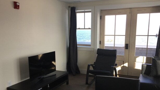 Ein Wohnzimmer mit einem Flachbildfernseher auf einem Standfuß, einem schwarzen Ledersessel und einer Couch. Zwei Fenster mit Vorhängen, eines davon mit Meerblick.