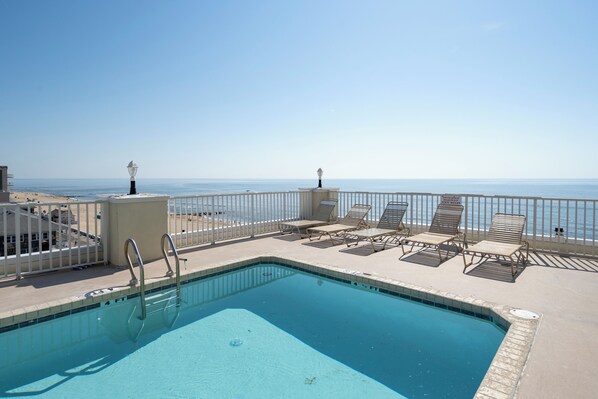 A pool with a view of the beach, surrounded by lounge chairs and a white fence.