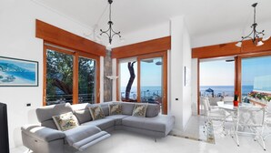Ein modernes Wohnzimmer mit einem grauen Ecksofa, gefliestem Boden, Holzbalkendecke und großen Fenstern mit Meerblick.