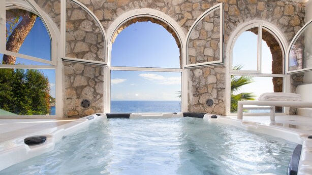 Ein Whirlpool mit Steinwänden und Bogenfenstern mit Blick aufs Meer.