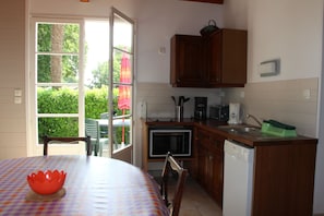 A compact kitchen with wooden cabinets, a microwave, a coffee maker, and a dishwasher. There is a table with a patterned tablecloth and a red bowl. A window with a view of greenery is visible. - 5 minutes from beaches country house with children's play area (Breuillet)