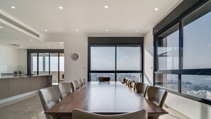 A conference room with a long table and chairs, large windows with a city view, and a modern kitchen in the background. - Jerusalem Penthouse (Jerusalem)