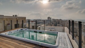 A rooftop hot tub with a wooden deck, surrounded by a metal railing, overlooking a cityscape with buildings and a bridge. - Jerusalem Penthouse (Jerusalem)