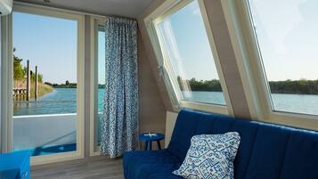 A room with a blue sofa, a patterned pillow, a small stool, and a view of a water body through large windows.
