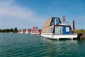 A row of houseboats with a wooden deck and a blue upper level, docked on calm water.