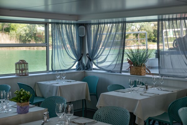 Un restaurante con mesas dispuestas para comer, vista al agua y una planta en el alféizar de la ventana.