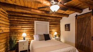A log cabin-style bedroom with a bed, two bedside tables with lamps, a ceiling fan, and a wooden door. - Fox Creek Cabin in the woods with stream (Narvon)