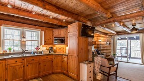 A rustic cabin interior with wooden floors, a kitchen with wooden cabinets, a dining area with a table and chairs, and a large window providing a view of the outdoors. - Fox Creek Cabin in the woods with stream (Narvon)