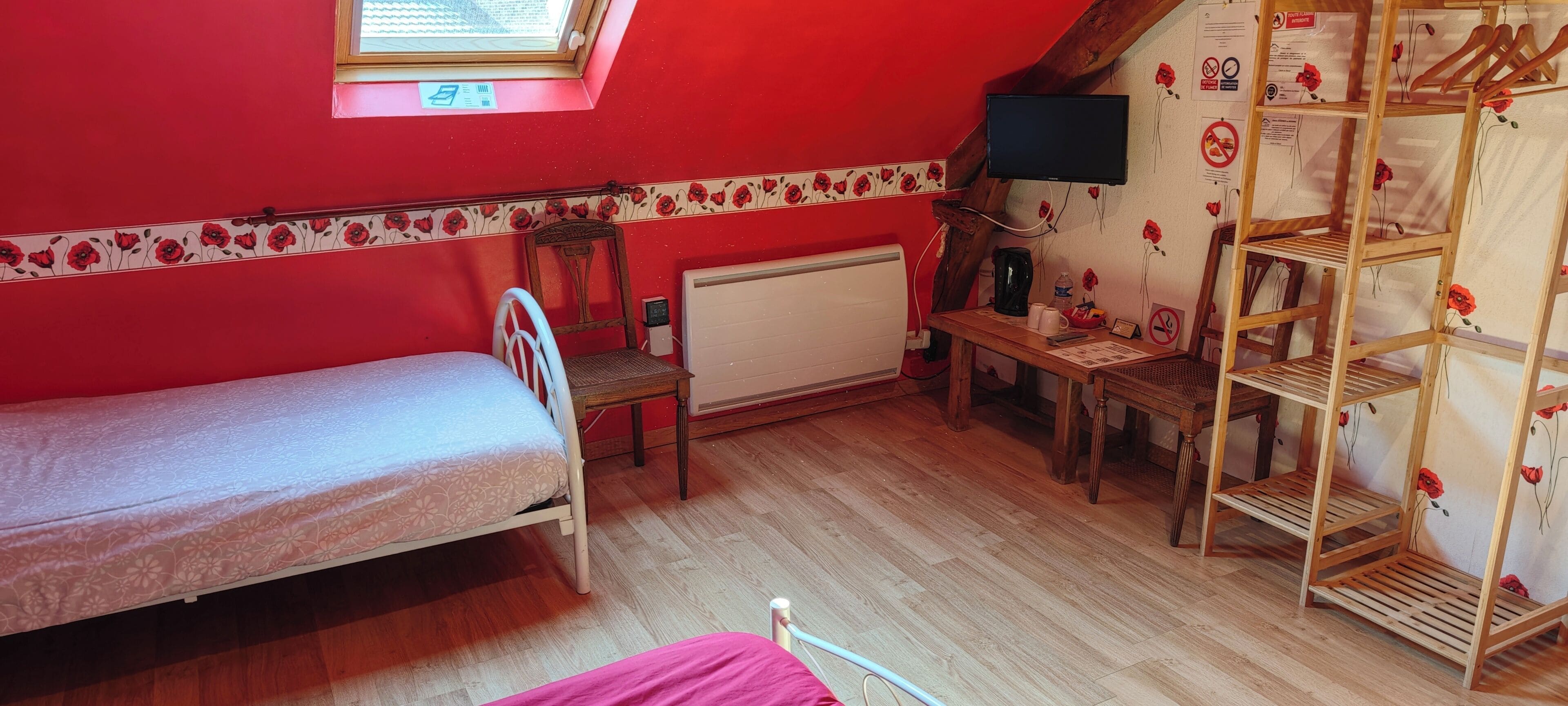 Ein kleines Zimmer mit einem Einzelbett, einem Holzstuhl, einem Heizkörper, einem an der Wand befestigten Fernseher und einer Holzregaleinheit mit Blumenmuster.