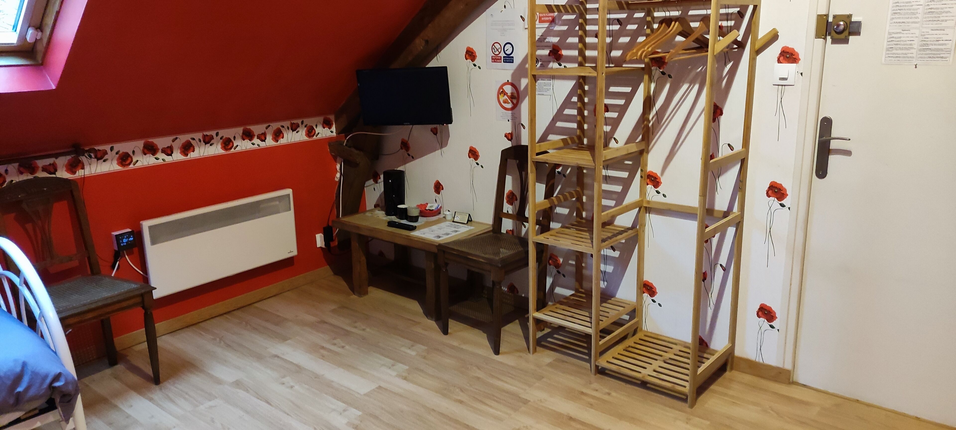 A room with a sloped ceiling, a wooden chair, a desk with a computer, a wooden ladder shelf, and a radiator.