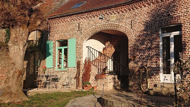 A brick building with a large arched doorway, green shutters, and outdoor seating. - Les Chambres du Relais (Liettres)