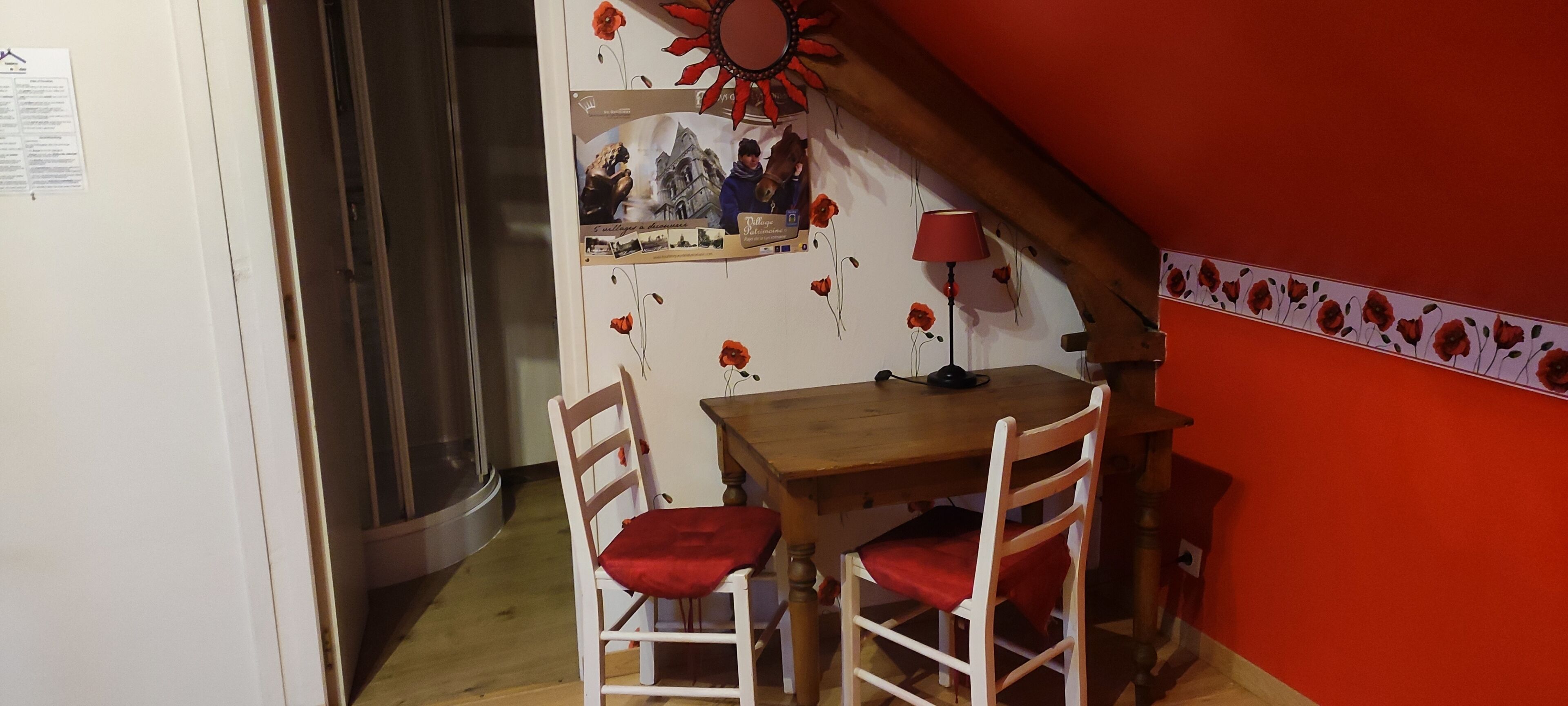 A small dining area with a wooden table and two chairs, a red lamp, and a wall decorated with red flowers and a vintage photo.