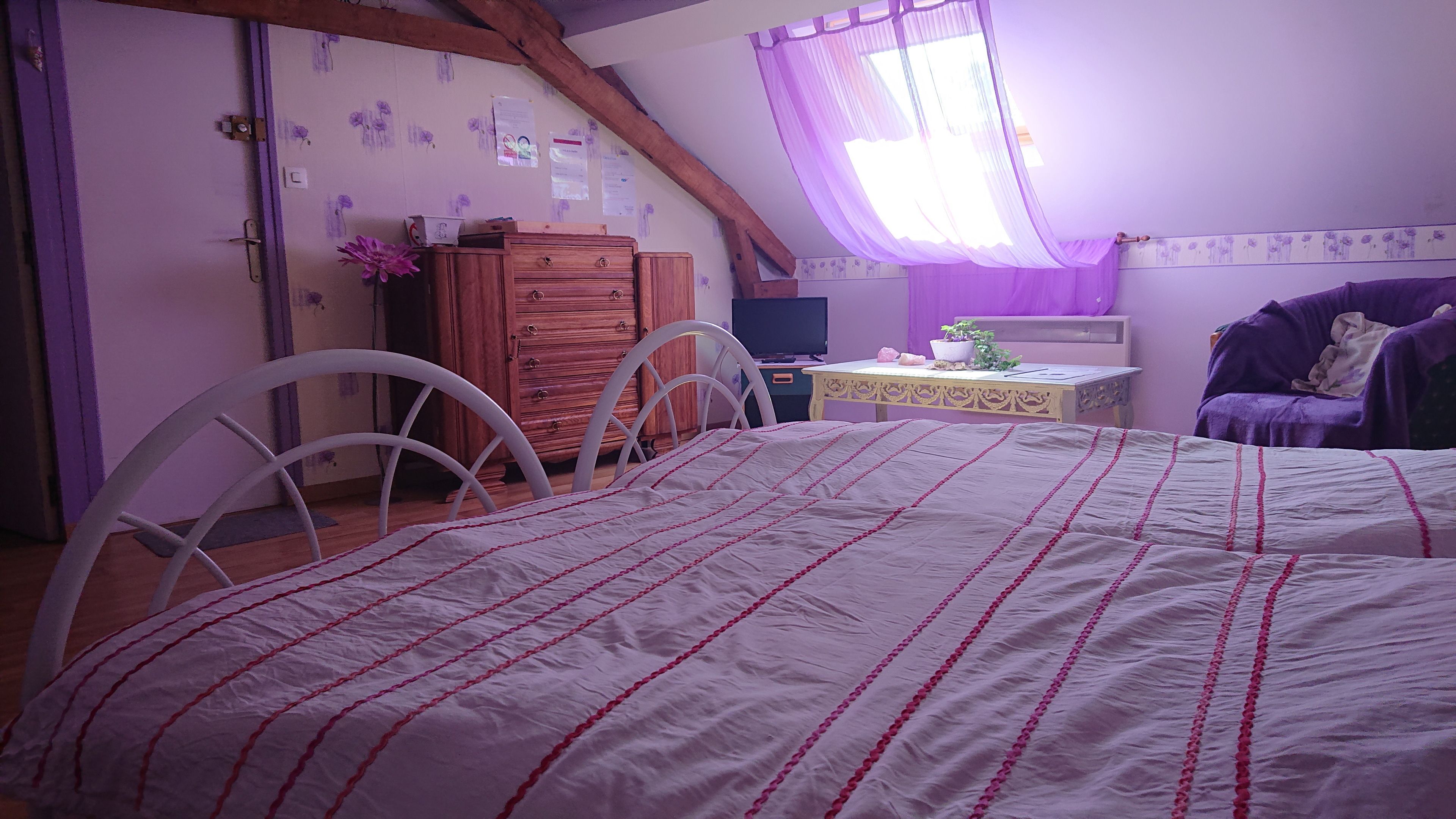 A bedroom with a bed, a dresser, a chair, a table, and a window with purple curtains.