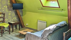A room with a sloped ceiling, a television mounted on the wall, a wooden chair with a green cushion, a small wooden table, and a sofa with a patterned cover. - Les Chambres du Relais (Liettres)