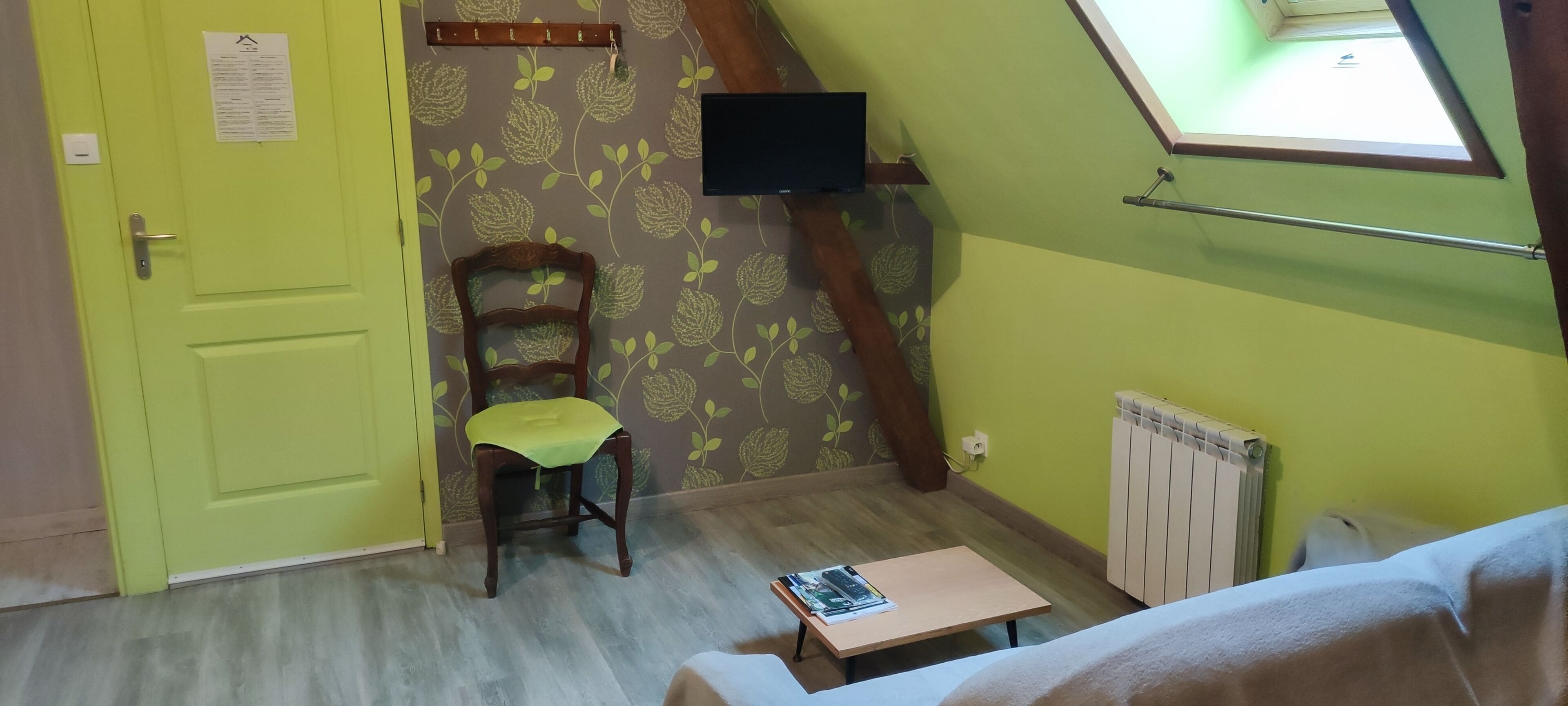 Una stanza con una porta verde, una sedia di legno con cuscino verde, un tavolino con delle riviste, un termosifone e una TV montata a parete.