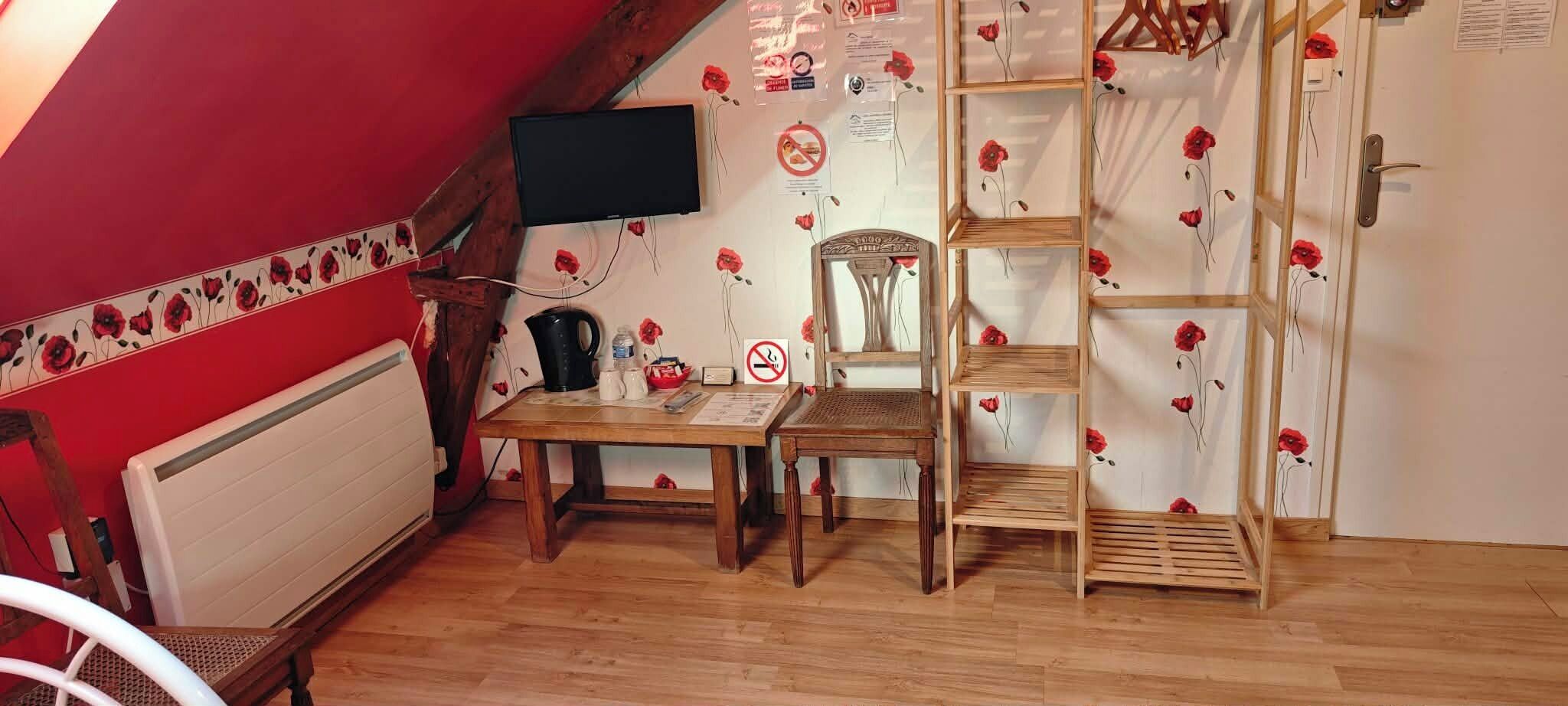 A room with a sloped ceiling, a wooden table with chairs, a black kettle, a white radiator, and a wall-mounted TV.