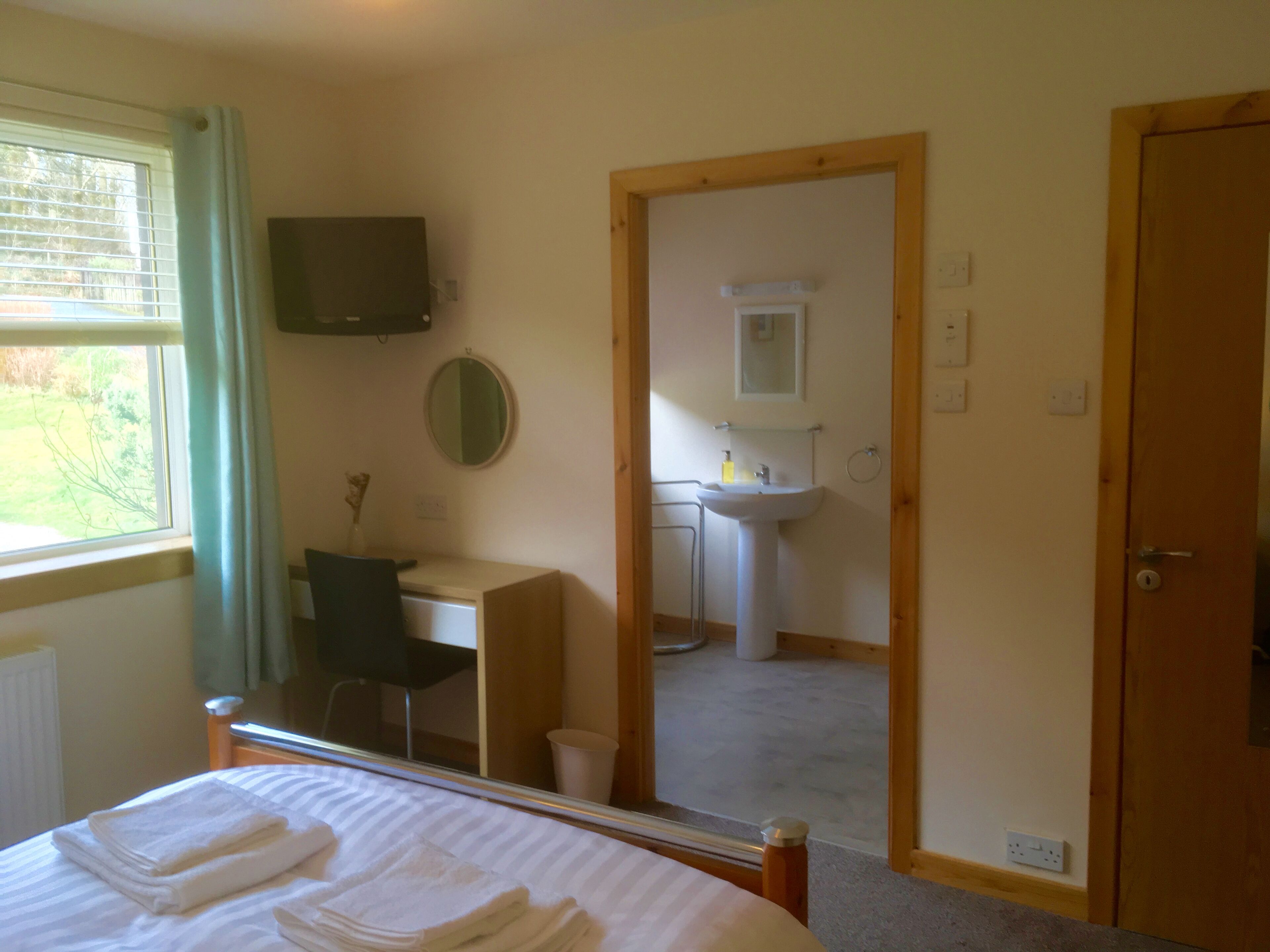 A bedroom with a bed, a desk, a chair, a mirror, and a bathroom visible through an open door.