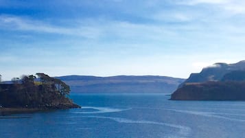 Un paysage côtier avec une mer calme, des collines lointaines et un petit bâtiment perché sur un promontoire rocheux.