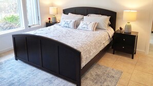 A bedroom with a dark wooden bed, white bedding, two bedside tables with lamps, and a view of greenery outside. - 6 Bedroom, 5 Bath Home with Pool, Sag Harbor - Near Bridgehampton (Sag Harbor)