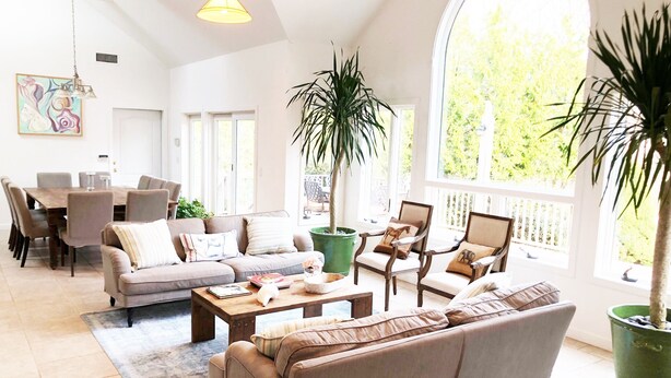 A spacious living room with a large sofa, a coffee table, and a dining area with chairs and a table. There are potted plants and a large window providing natural light. - 6 Bedroom, 5 Bath Home with Pool, Sag Harbor - Near Bridgehampton (Sag Harbor)